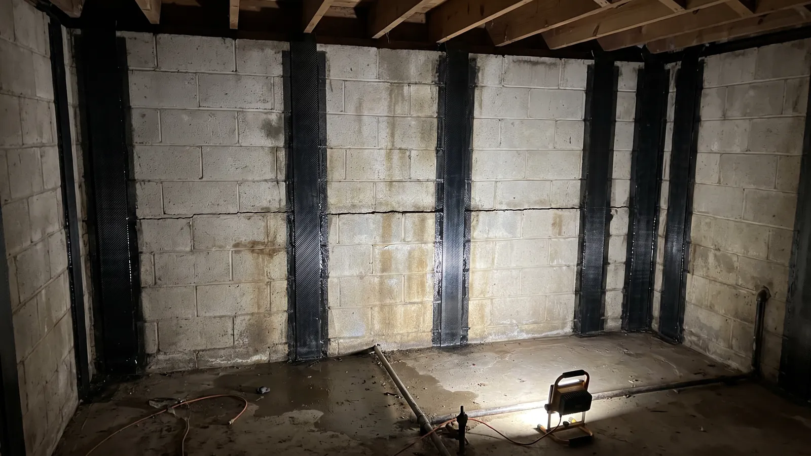 Vertical carbon-fiber reinforcement straps epoxied to a concrete block basement wall that shows a horizontal crack and slight inward bow, in an unfinished Alabama basement